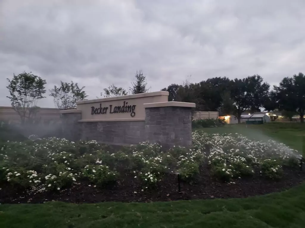 Sprinkler system watering flower beds near the 'Becker Landing' entrance sign.