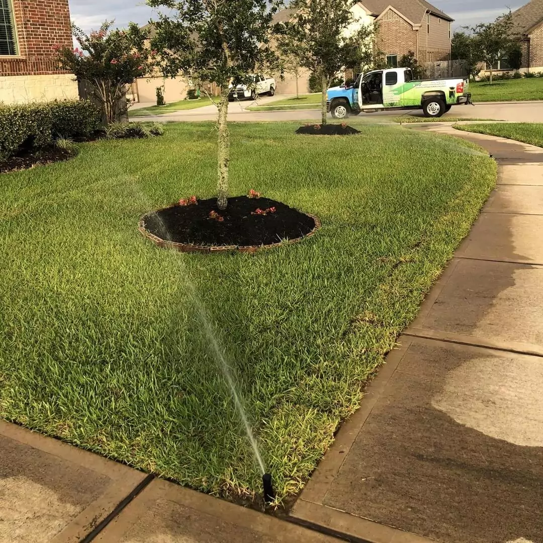 A yard with sprinklers active, spraying water near a tree surrounded by mulch, with a Murf Turf Sprinklers & Water Management truck parked in the background.