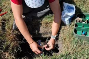 Person performing maintenance on an underground sprinkler system, working with wires and tools in a grassy yard.