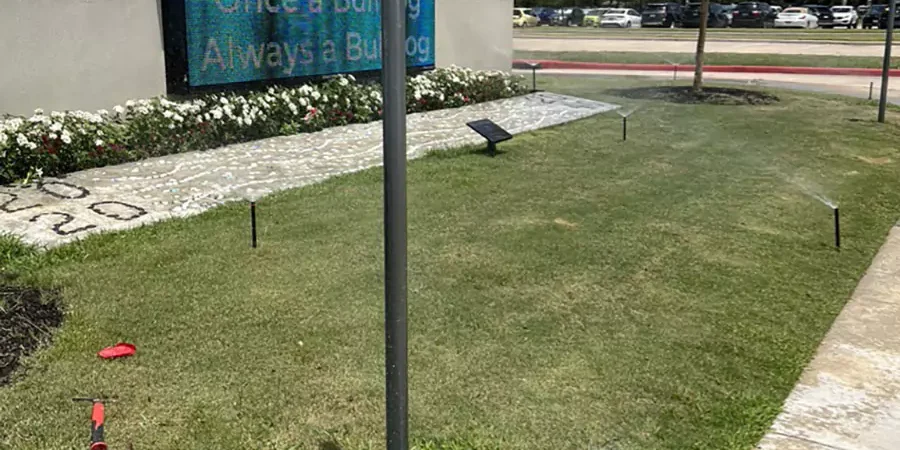 Lawn sprinkler system operating near a school sign with flower beds and green grass