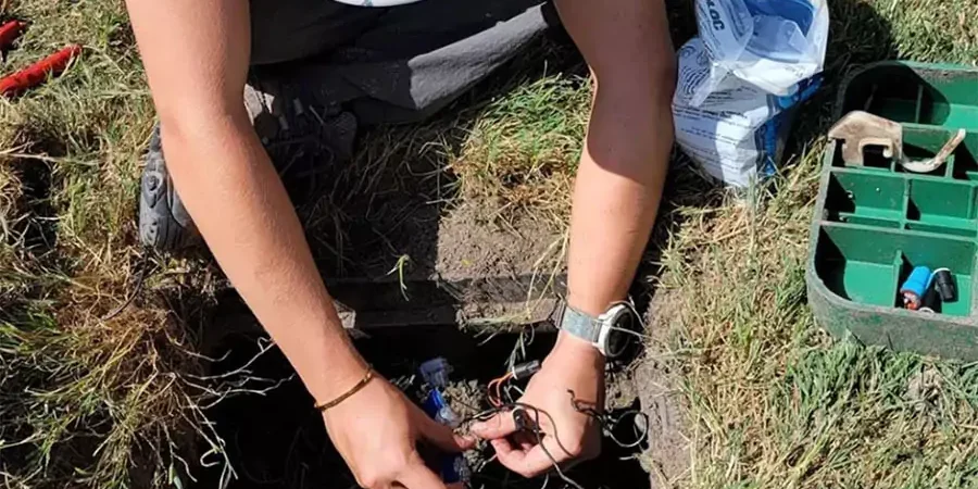Person performing maintenance on an underground sprinkler system, working with wires and tools in a grassy yard.