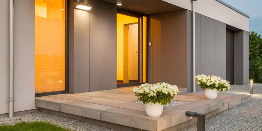 Modern home entrance with warm lighting, potted white flowers on the porch, and sleek outdoor lighting fixtures.